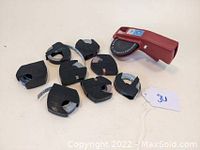 Vintage red Dymo label maker with dial and lever, shown with seven black plastic label tape cartridges containing adhesive tape rolls.