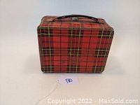 Front view of vintage steel lunch box with red plaid pattern, black handle, and metal latch, showing slight wear and scratches