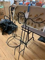 Two black metal garden hose holders with a decorative metal wall piece behind them on a wooden floor.