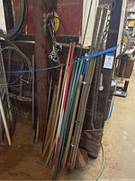 Photo of various long-handled farm and garden tools leaning against a wooden wall with metal wiring coils hanging above.