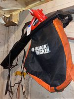 Black and Decker leaf blower with orange and black fabric collection bag against a wall.