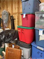 Stacked Christmas decor storage bins in various colors with labels and some visible contents such as artificial poinsettias.