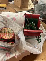 Photo of red metal Christmas sleigh, green cushioned seat, holly and bells decoration, and three Christmas patterned baskets nearby on floral fabric surface.