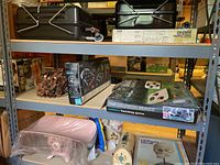 Two Coleman two burner propane stoves displayed on shelving, one in box, other uncovered. Thanksgiving pinecone centerpiece and beanbag yard game boxed set also shown, along with smaller classroom items on lower shelf.
