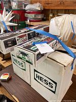 Photo showing three Hess trucks in original packaging, two smaller boxes on top of a large white box with 'Hess' printed on them. Packaged with blue painter's tape securing boxes.
