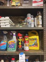 Photo of household cleaning and maintenance supplies on metal shelves including spray cleaners, insecticides, coolant, primers, and assorted jars.