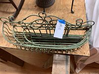 Green metal window box planter featuring decorative scrollwork, photographed on wooden surface with tag