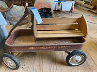Photo showing the full view of the vintage red metal wagon labeled 'Radio Astronaut' with attached wooden bookshelf above it. Wagon has four rubber tires with white metal rims, wooden shelf is mounted to the wagon.
