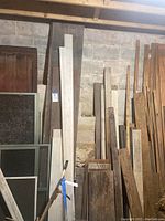 Stacks of assorted scrap wood planks and boards leaning against concrete block wall. Various sizes, both narrow and wide pieces visible.