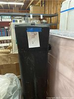Tall black fabric carrying case for pop-up tent or canopy with wheeled base shown standing upright in storage or warehouse setting.