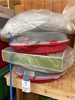 Stack of round plastic wreath storage containers with red locking lids. Plastic bags and protective packaging covering the wreaths inside. Visible tags with handwritten notes describing contents and condition.