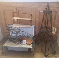 Multiple assorted household items including laptop monitor riser with drawer, desk shelves organizer, wire fruit basket, wall hanging shelves, and Eiffel Tower wine rack, placed against a wooden cabinet.