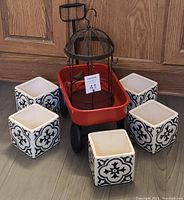 Red mini wagon with black wheels holding a rustic metal bird cage surrounded by six white square ceramic planters featuring black decorative patterns