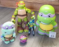 Overview of the lot featuring five Teenage Mutant Ninja Turtles items: large Donatello action figure, mini Leonardo action figure, Donatello plush, ceramic Donatello bank, and small themed ball arranged on floor.
