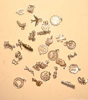 Collection of various vintage sterling silver bracelet charms in assorted shapes including a butterfly, violin, airplane, baby stroller, crown, letters, and other motifs scattered on a flat surface.