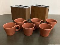 Photo showing two vintage mid-century modern style kitchen canisters with wood paneling and chrome lids, along with six reddish-brown terracotta ceramic cups with handles.