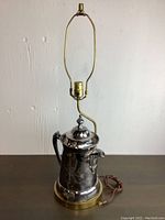 Full view of the vintage silverplated teapot lamp showing the lamp fitting, brass-colored base, and teapot body with cord.