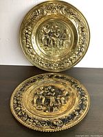 Photo showing both brass plates with intricate stamped scenes and floral borders set on table and wall