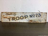Photo of the full metal sign leaning against a white wall showing the entire text and condition of the sign with rust and wear clearly visible.