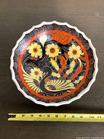 Overhead shot showing the vibrant floral and bird pattern inside the ceramic bowl with scalloped edges and a tape measure below for scale showing approximately 10 inches diameter.