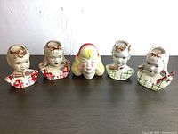 Five vintage ceramic wall-mounted heads displayed on a wooden surface. Includes four children's busts with plaid shirts in red or green and a central female head with blonde hair. Each piece shows some age wear and glazing imperfections.