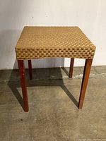 Front view of square woven wicker top side table with wooden legs on concrete floor against white wall.