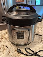 Front view of Instant Pot showing overall appliance with lid closed, power cord attached, stainless steel exterior, and black top handle.