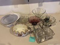 Various glass bowls, including a clear fluted bowl, a pink glass bowl, a square divided glass utensil holder with metal base, shot glasses, and stacked silver toned chargers.
