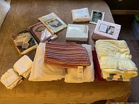 Photo showing assorted folded bath towels in white, red striped, cream, and packaged linens including tablecloths and sheets.