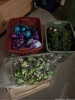 Top view of storage bins showing multiple purple and blue Christmas ornaments and a bag of green ornaments with artificial evergreen garland tote