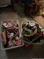 Two white plastic totes, one with assorted Christmas ribbons, gift boxes, and tags; other with Christmas wreath, garland, and decorative items on concrete floor.