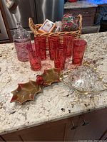 Photo showing six cranberry glass drinking glasses, cranberry glass candy jar with lid, vintage cardinal nut dish, glass Christmas tree serving dish, wicker basket with soaps and potpourri on granite countertop.