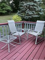 Two reclining deck chairs and two small side tables on a red wooden deck outdoors with greenery in background