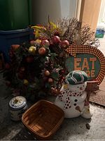 Photo of grapevine wreaths decorated with faux apples and leaves, snowman cookie jar, Longaberger basket, and tin 'Eat Here' sign arranged together on floor with some packing bins in background.