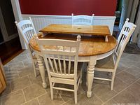 Wood kitchen table with natural wood top and white legs, surrounded by 4 white wooden chairs with upholstered seats on a tiled floor.