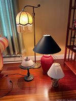Four assorted lamps including a tall floor lamp with beige fabric shade and three table lamps of various sizes and colors, arranged on a wooden floor.