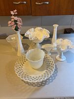 Full view of all milk glass items on kitchen counter showing variety of vases, footed bowls, mini planter, basket piece, and lattice-edged platter.