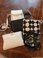 Five purses arranged on a wooden surface showing diversity in style and materials: two leather crossbody purses (black Ann Taylor, white), vintage Walborg clutch with chain, black and white patterned Mad for Design bag, and white woven bag.