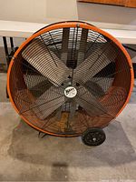 Front photo of the Maxx Air floor fan showing metal blades behind a black protective grille and orange housing rim with two wheels.
