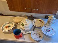 Overview of porcelain and ceramic servingware items including cake plate, soup tureen, pitchers, pans and bowls with floral and checkered patterns