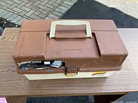 Closed brown and beige plastic Plano fishing tackle box on a table, showing signs of use and a label on the front.