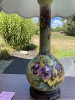 Close-up photo of the first vintage table lamp showing the vase-shaped ceramic base painted with purple, yellow, and white pansy flowers on a light yellow background with a scalloped linen shade.
