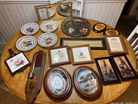 Full assortment of decorative plates, plaques, framed art displayed on round wooden table under bright lighting, showing variety and condition.