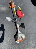 Overall front view of Stihl weed eater lying on asphalt with red gas can, 2 stroke oil, and replacement string nearby