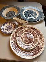 View showing assortment of blue and brown willow ware plates, small mini sauce boat, and purple floral patterned bowl on table.