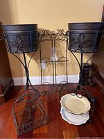 Photo showing full set of wrought iron decorative items arranged on floor including two metal buckets with wrought iron stands, double tier server with pie plates, wrought iron basket, plate hangers, and book stand.