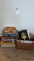 Stack of various hardcover and paperback books next to an open box containing brass door knockers with a cardboard floor background.