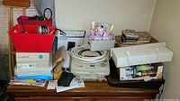 Wide view of lot including red storage containers with binocular box and IP camera, Panasonic fax machine, boxes, notebooks, wires, and art supplies