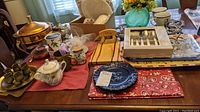 Overview of table with copper chafing dish, ceramic serving platters, plates, mugs, pewter shot glasses, flatware in box, chopping set, decorative green glass vase, and Lenox sugar bowl.