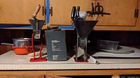 Two manual food grinders, plastic trays, food mill and enamel bowls on countertop under cabinets.